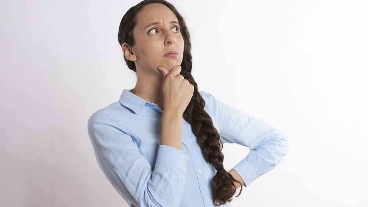 A woman with a long dark braid and a light blue button shirt stands against a plain white background, one hand under her chin and the other on her hip, looking up and to the side with a furrowed brow.