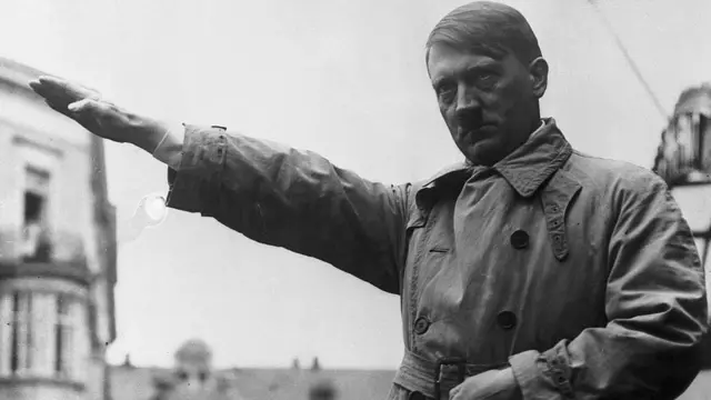 A black and white photograph of Adolf Hitler in a belted trench coat with his right arm extended, buildings behind him.