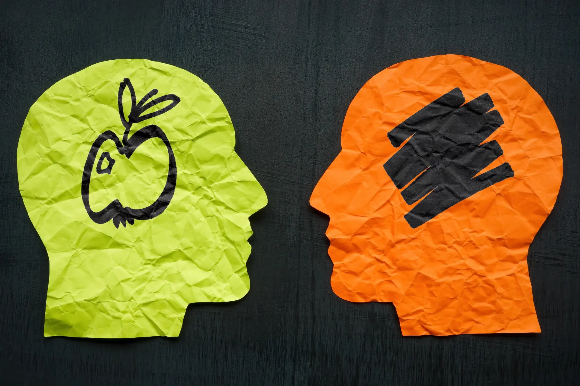 Two paper head silhouettes on black, one green with a drawn apple, one orange with scribbles.