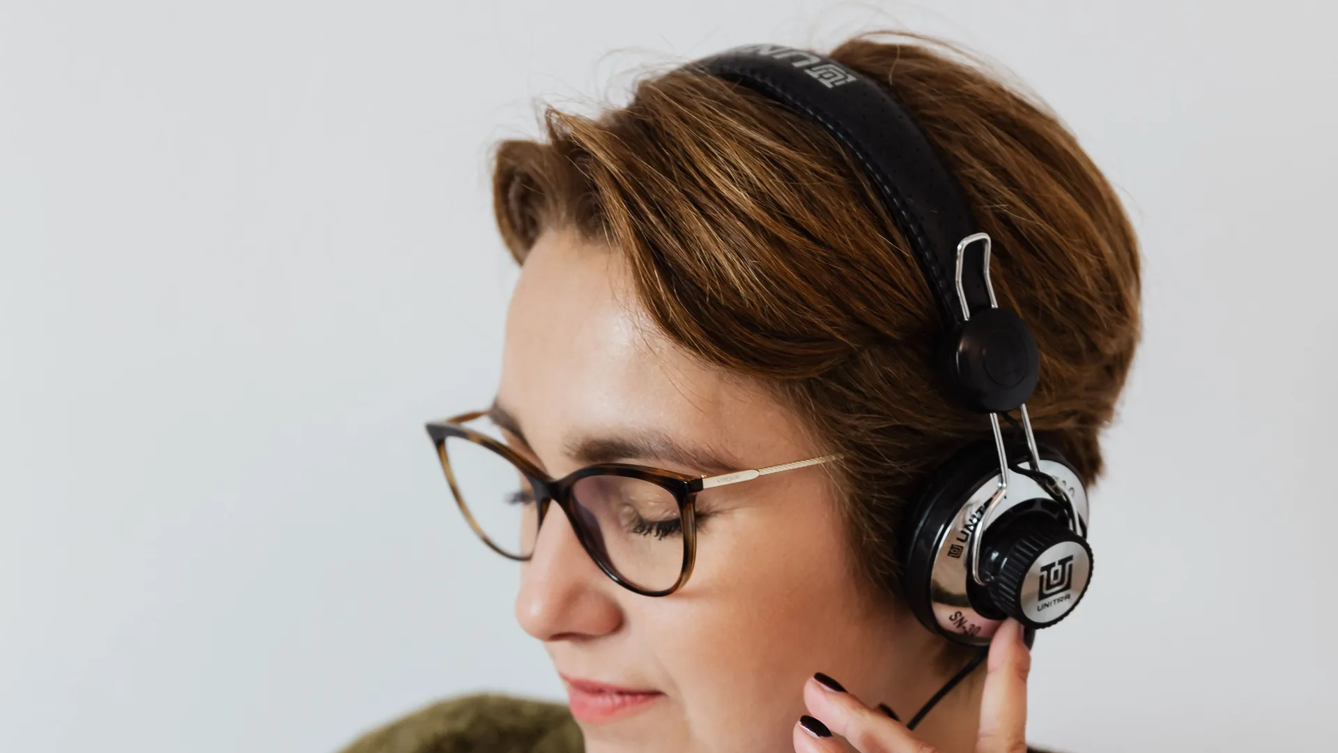 A woman with glasses and over-ear headphones listens with her eyes lowered and one hand at the cable.