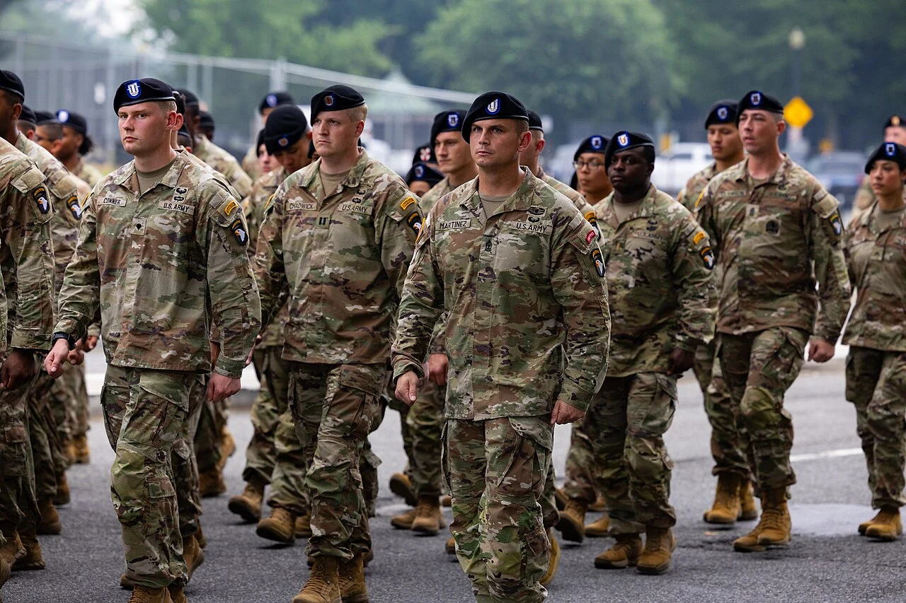 Soldiers in camouflage uniforms and black berets march in ranks along a paved road in daylight, with name tapes reading U.S. Army and blurred trees and vehicles behind them.