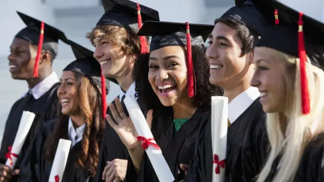 Six graduates in black caps and gowns with red tassels standing in a row outdoors, smiling and holding rolled diplomas tied with red ribbon.