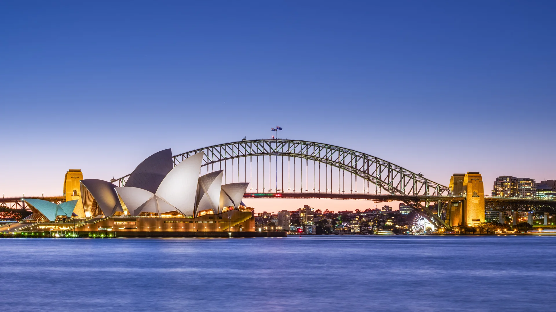 The white sail shaped roofs of the Sydney Opera House lit at dusk in front of the steel arch of the Sydney Harbour Bridge, with city lights across the water and a clear sky fading from deep blue to orange at the horizon.
