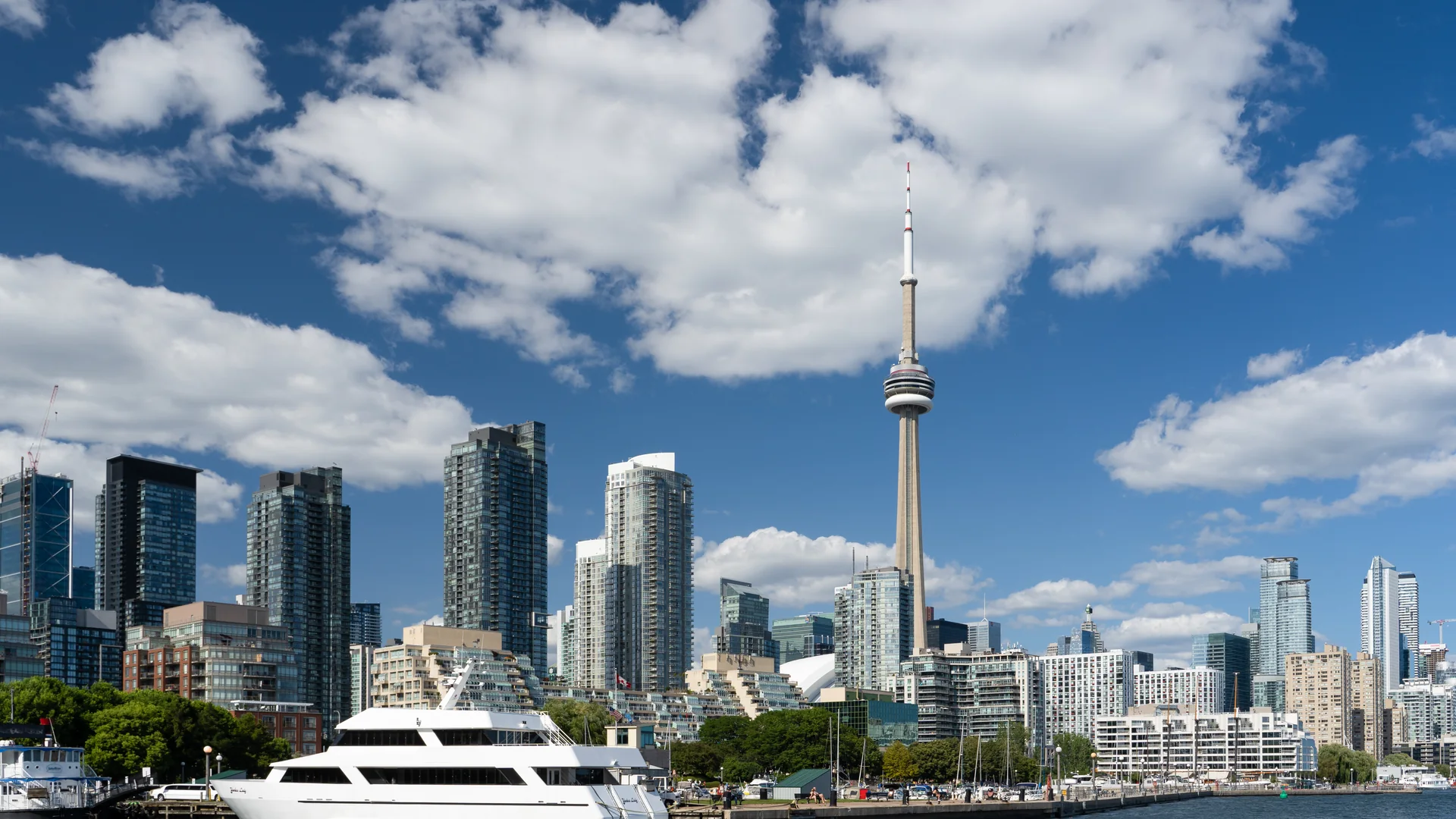 The Toronto skyline seen across the water from Ireland Park on a sunny day, with the CN Tower rising above glass condominium towers, cumulus clouds overhead and a white yacht moored in the foreground.
