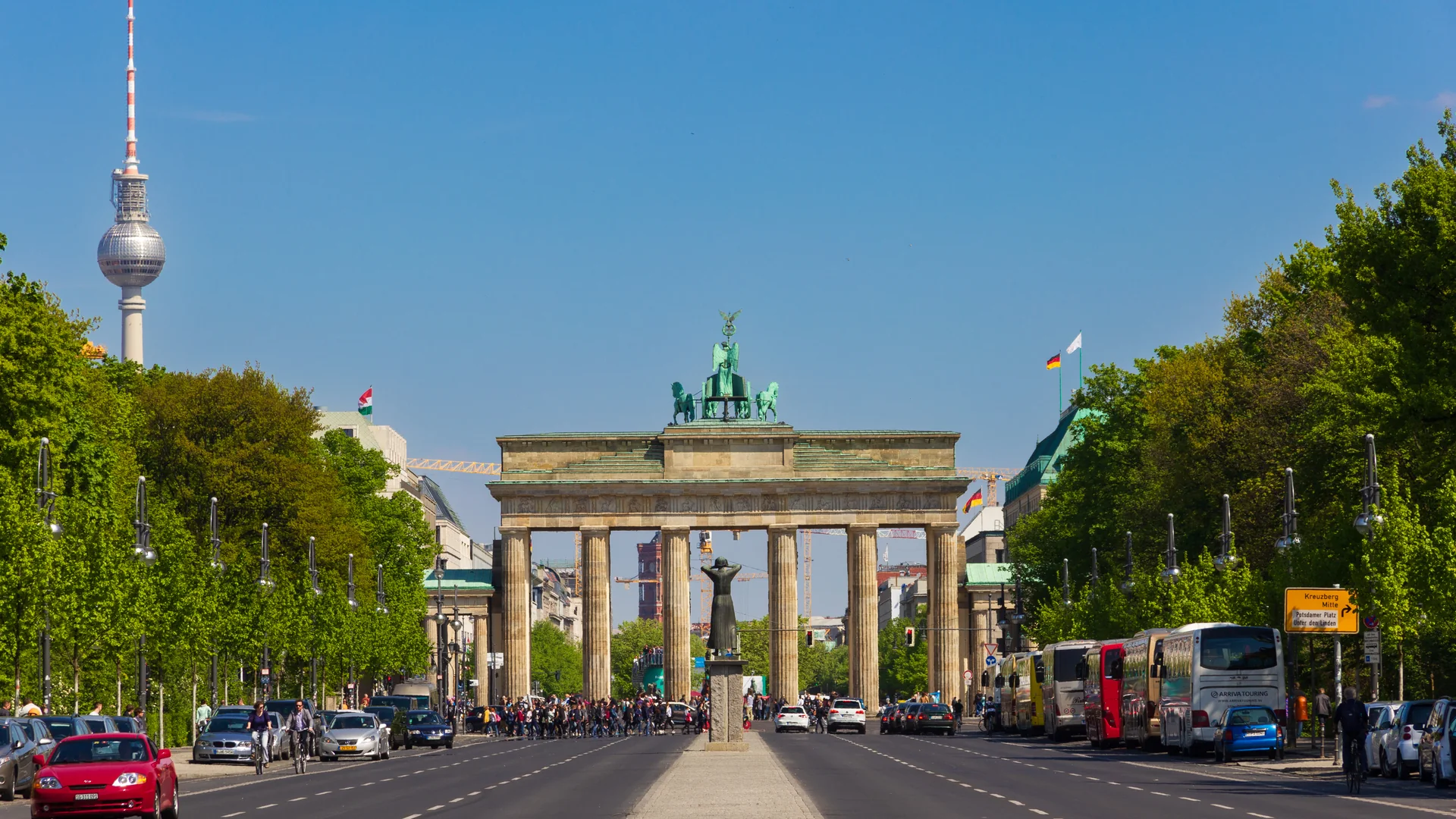 A wide, straight avenue lined with green trees leads toward the Brandenburg Gate in Berlin, with the bronze quadriga on top of the gate, the television tower rising at the left, cars and coaches parked along the roadside and a crowd of pedestrians crossing in front of the gate under a clear blue sky.