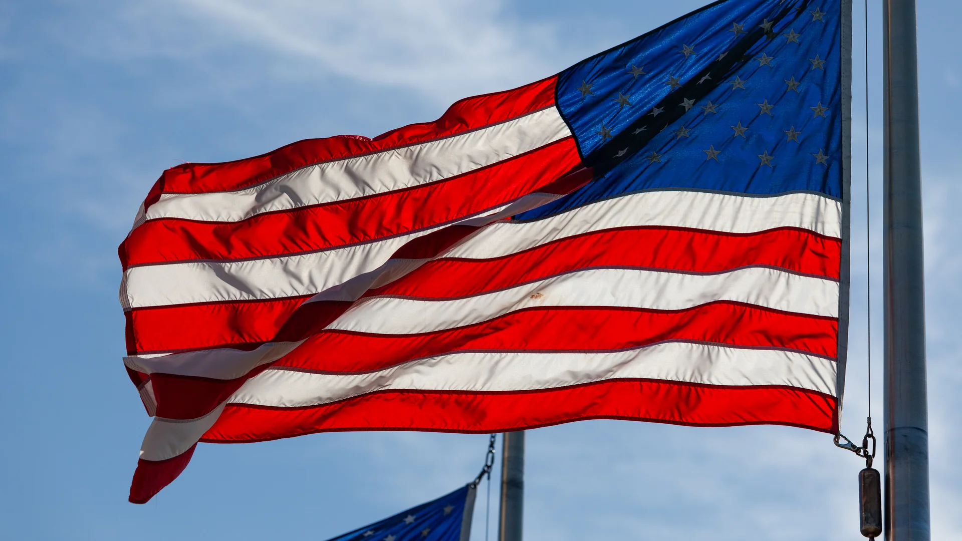 A close up of an American flag waving against a blue sky with thin clouds, its stars and red and white stripes lit by the sun, with a second flag partly visible behind it on a lower pole.