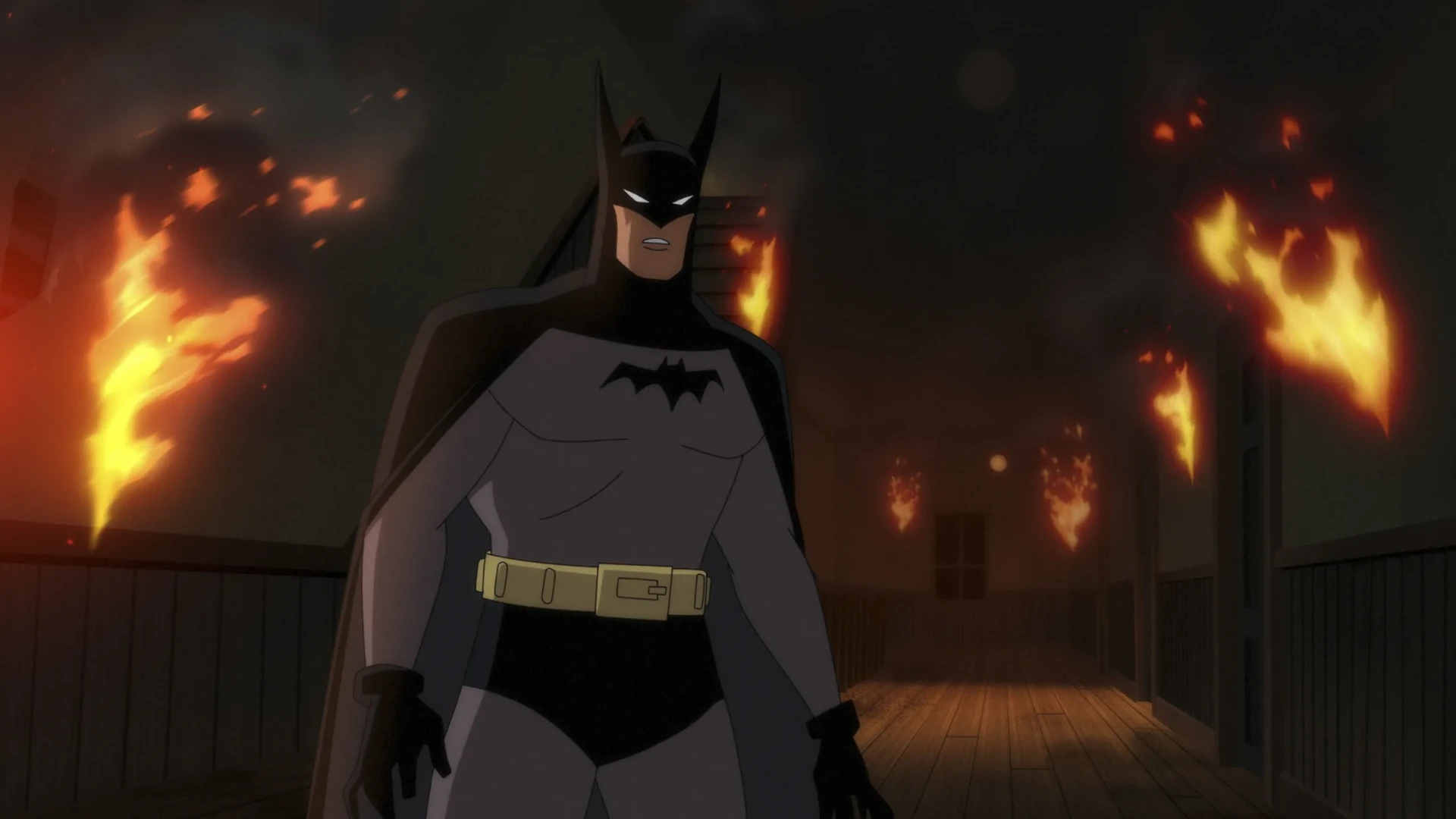 Animated Batman in a gray suit with a black cape, cowl and gold utility belt, standing in a wooden corridor with flames burning along the walls and dark smoke behind him.