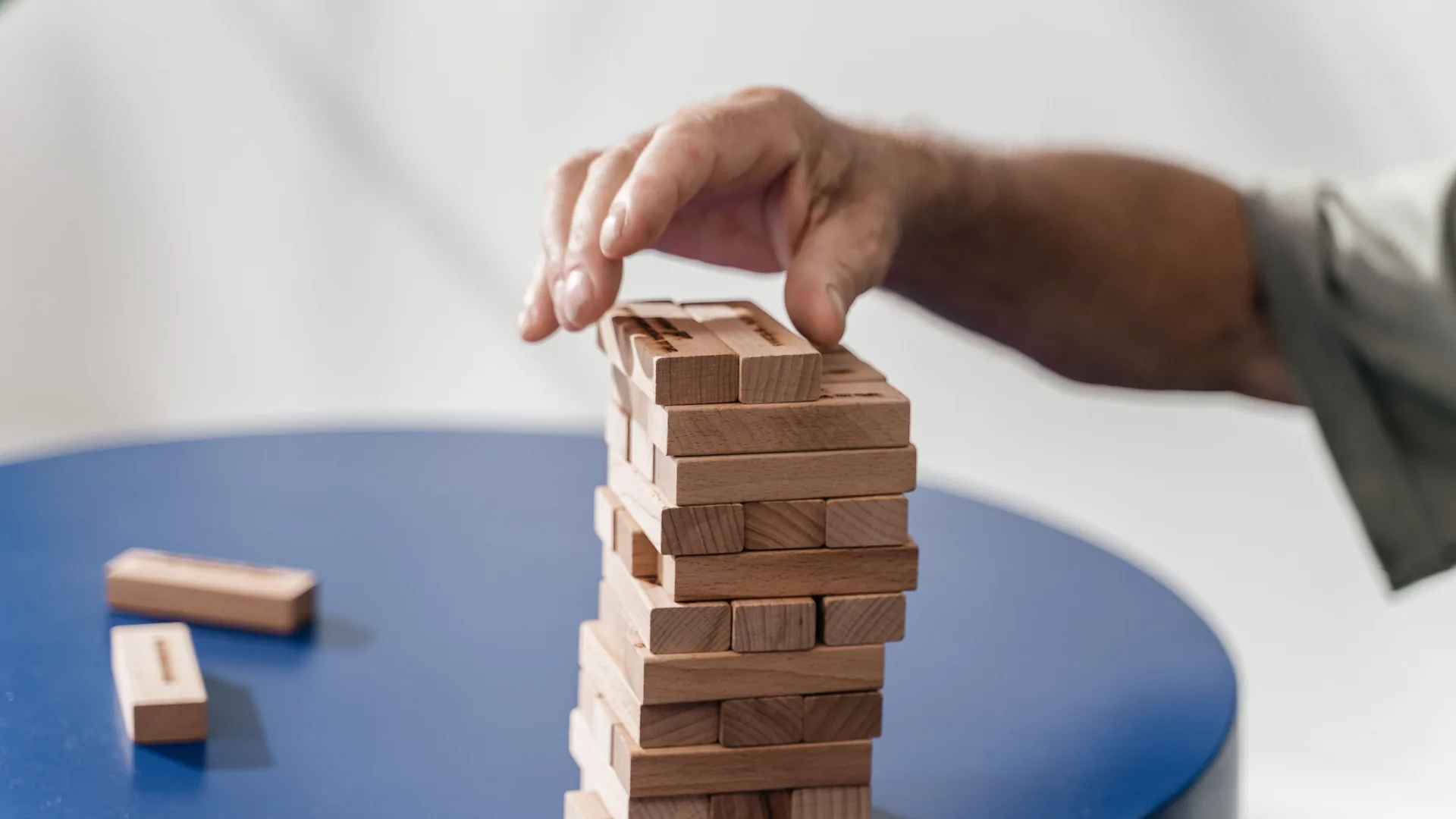 A hand places a wooden block on top of a stacked block tower on a blue table.