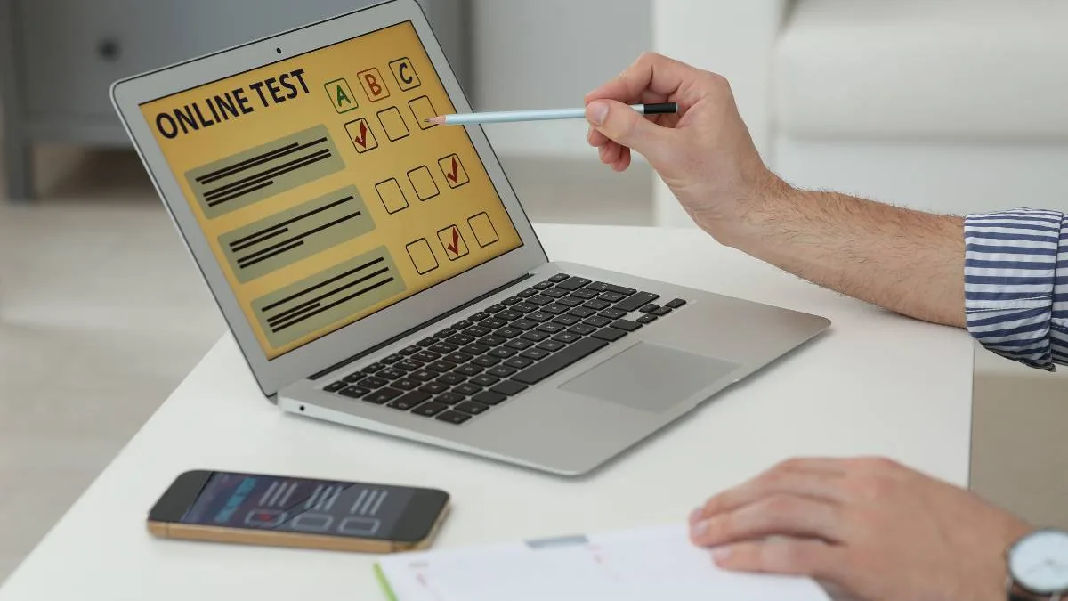 A hand holds a pencil toward a laptop screen showing a yellow form headed ONLINE TEST with answer boxes marked A, B and C and several ticked boxes, beside a smartphone and an open notebook on a white table.