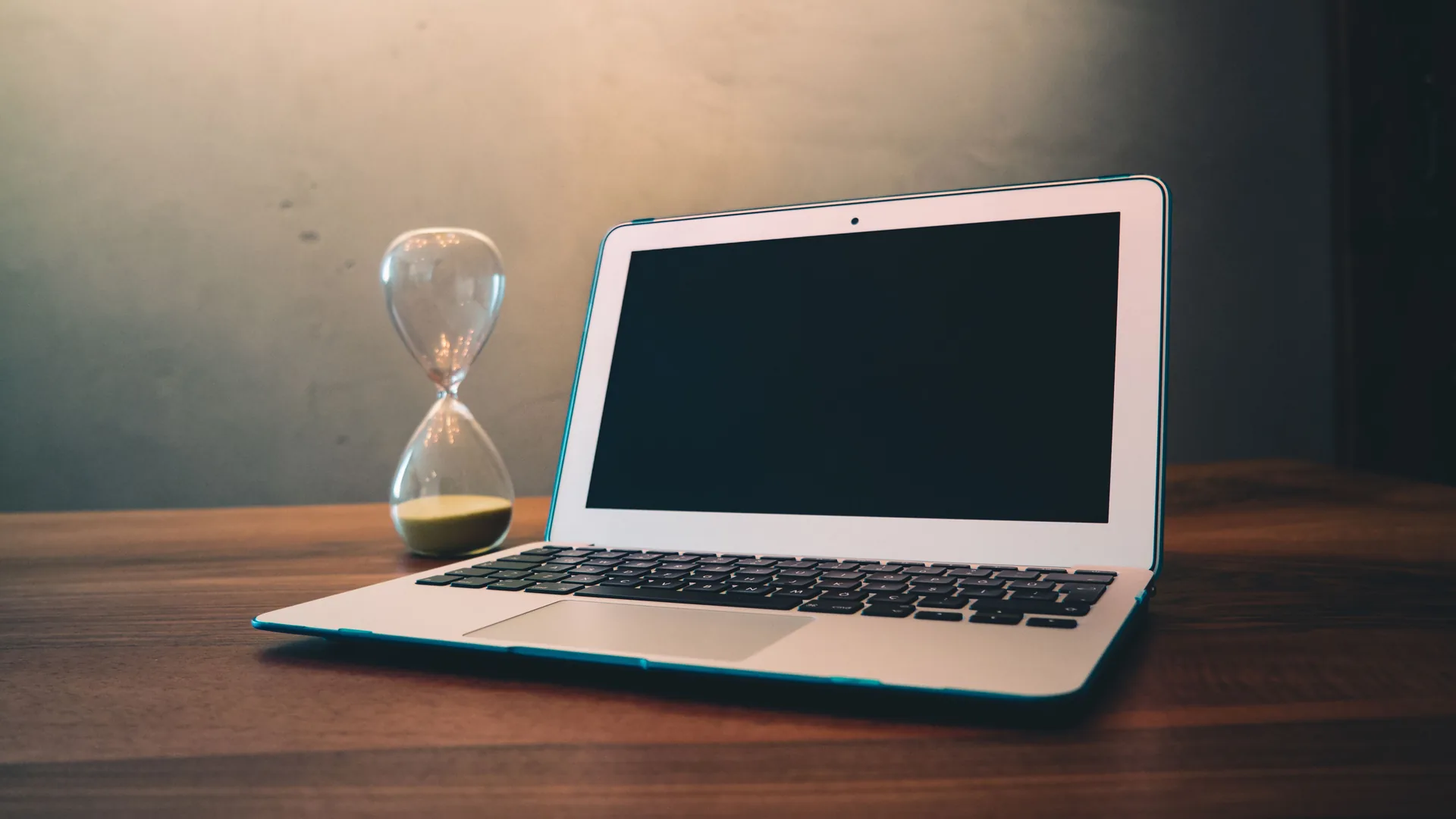 A laptop with a dark screen sits on a wooden table next to an hourglass.