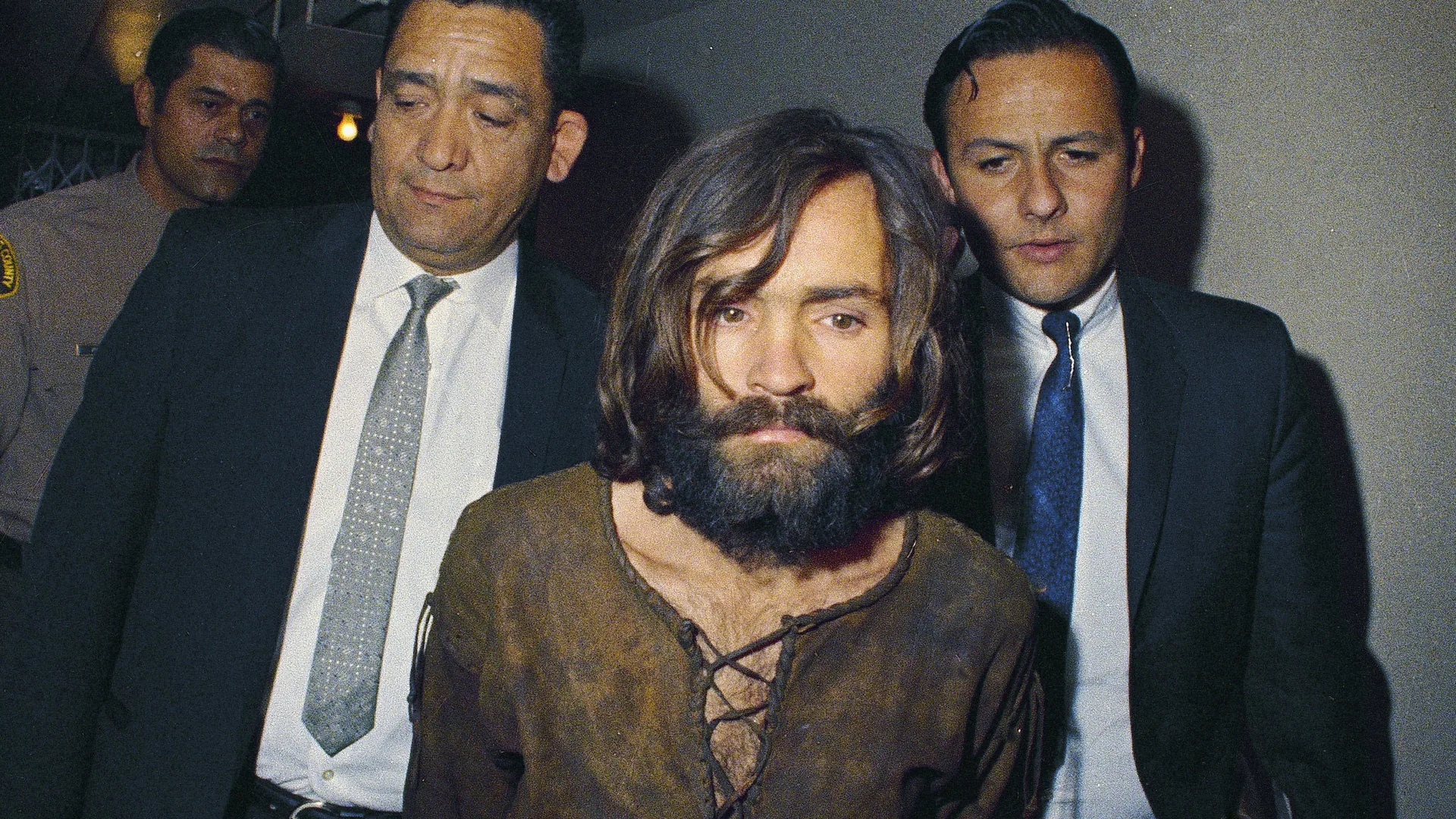 Color photograph of a man with shoulder length brown hair and a full dark beard, wearing a brown shirt laced at the neck, walking between two men in dark suits and ties who look downward, with a man in a tan uniform partly visible behind them.