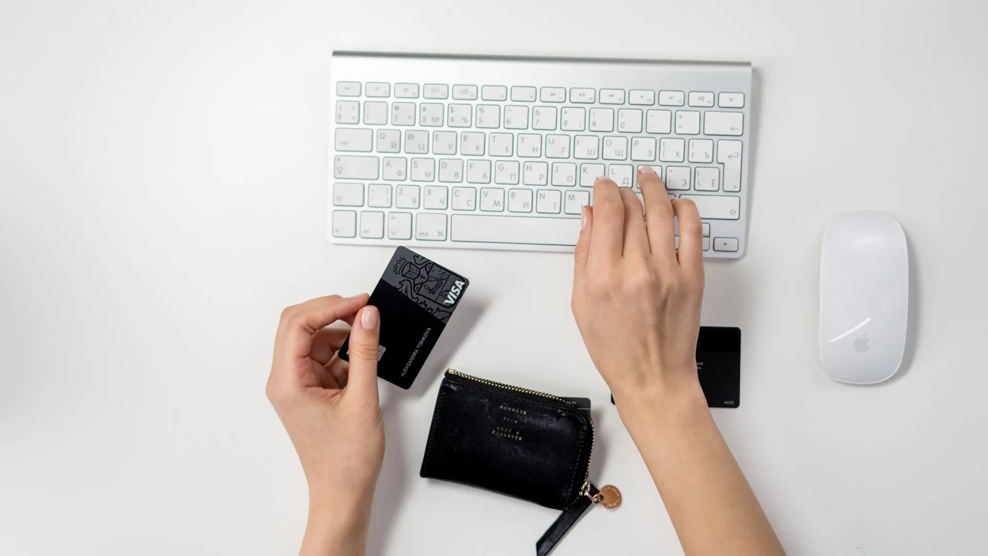 A top view of a white desk where one hand holds a black Visa card above a small black zip wallet while the other hand types on a white wireless keyboard beside a white mouse.