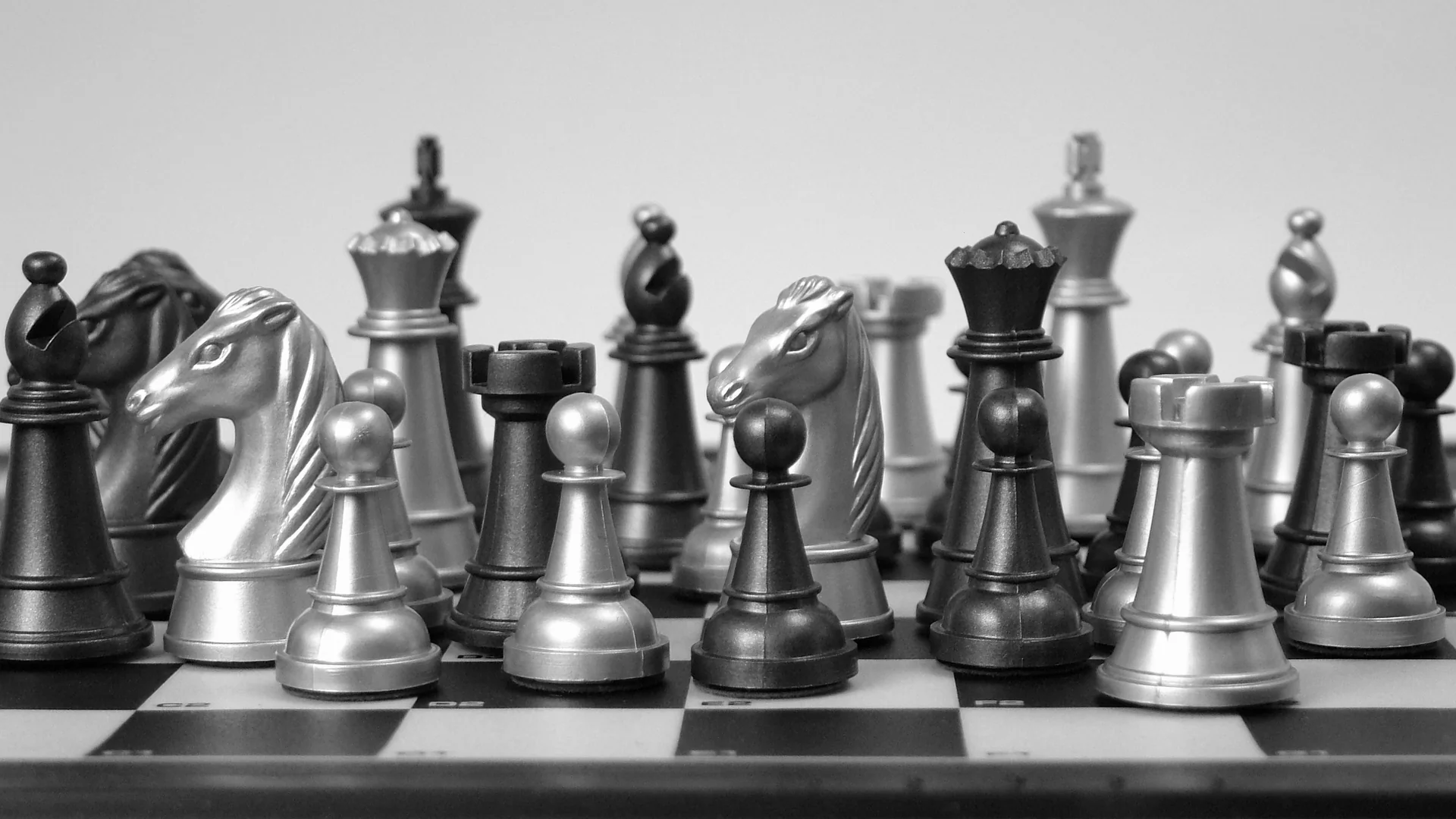 Silver and dark chess pieces stand close together on a board with printed square coordinates, with knights and pawns in the foreground against a pale background.