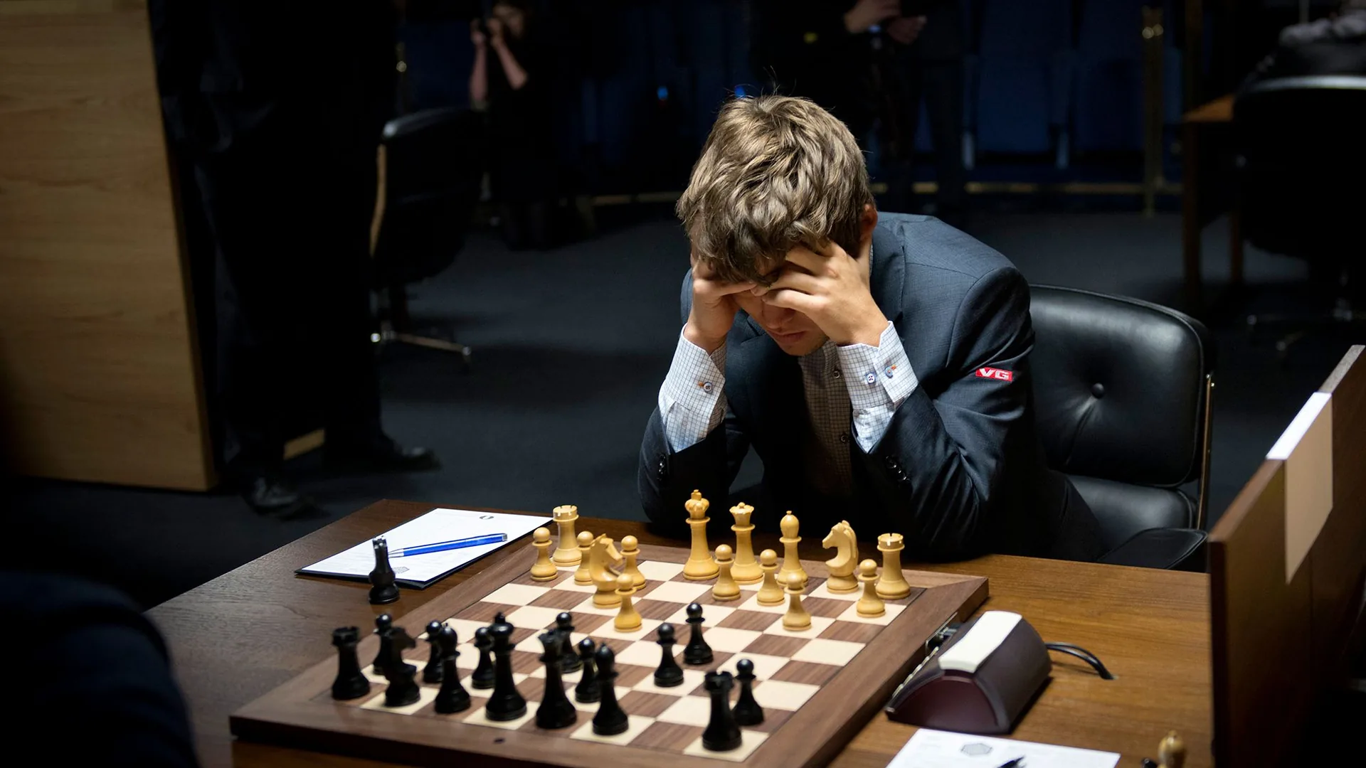 A young man with light brown hair sits at a wooden table with his head held in both hands, looking down at a chess board with the pieces set up mid game, a chess clock and a score sheet with a blue pen beside the board and a photographer standing in the dark background.