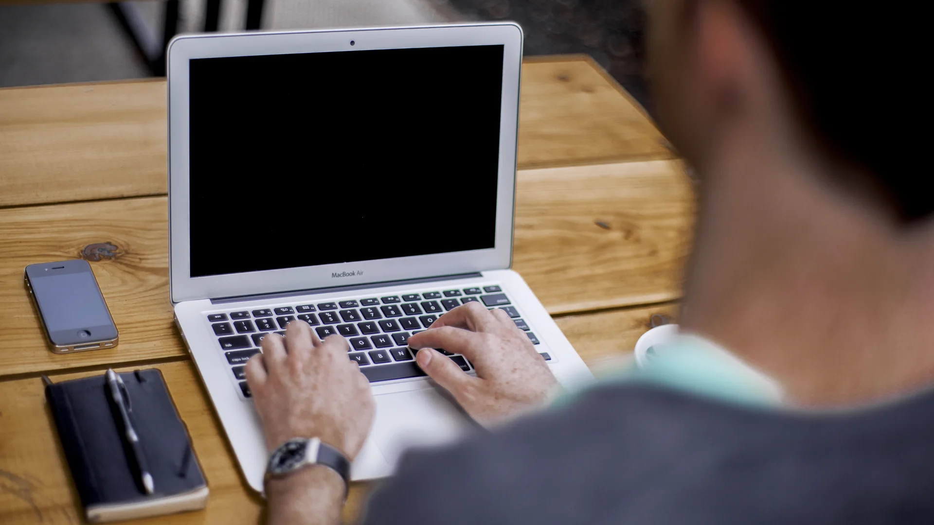 A man seen from behind types on a silver laptop with a dark screen at a wooden table, with a smartphone to his left, a black notebook with a pen beside it, and a watch on his wrist.