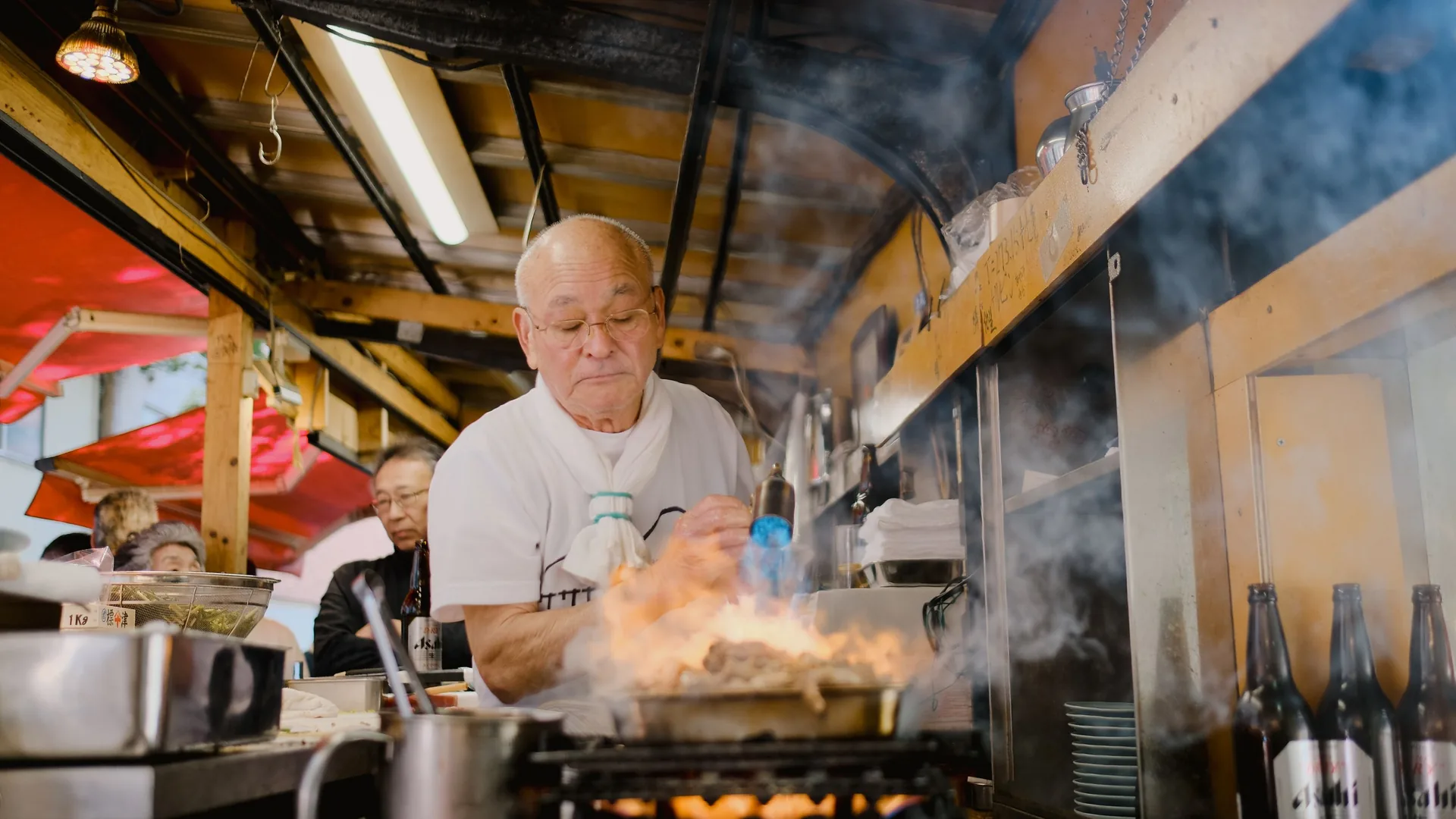 A cook works over a flaming pan in a busy restaurant kitchen, with other cooks behind him.