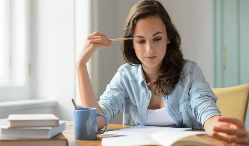 A woman at a desk with a pencil held to her temple, looking down at a page of notes beside a blue mug and a stack of books.