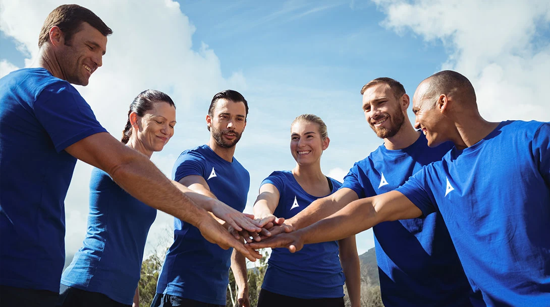 Six people in matching blue shirts, several with a small white triangle emblem, stand in a circle outdoors, smiling, each reaching one hand into the center so the hands are stacked together, photographed from below against a blue sky with scattered white clouds.