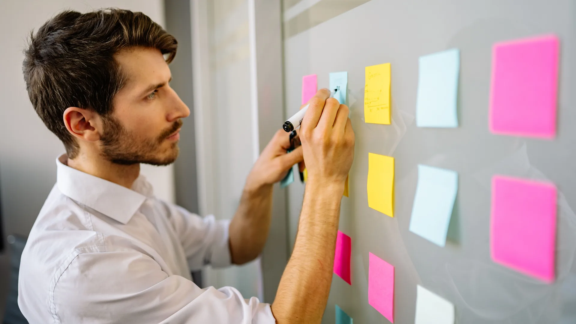 A man in a white shirt writes on a sticky note on a glass wall covered with pink, yellow and blue notes.