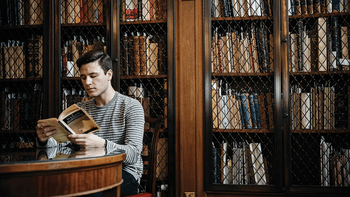 A man reads an old book at a round table in front of tall wooden bookcases with metal grilles.