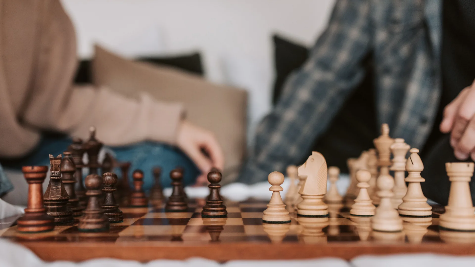 Two people sit at a wooden chessboard with the pieces in play between them.