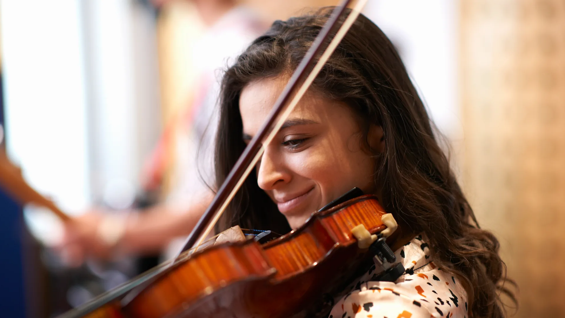 A woman with long dark wavy hair and a patterned blouse smiles with her eyes lowered as she plays a violin, the bow crossing in front of her face, with blurred people and warm light behind her.