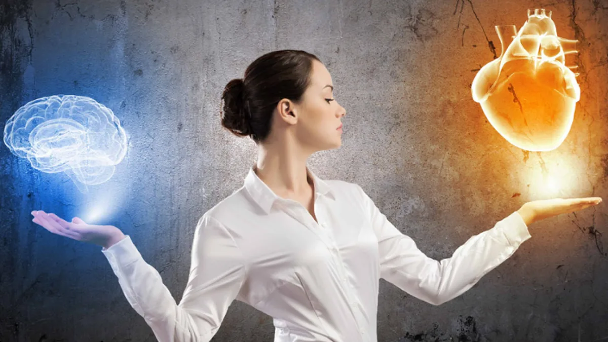 A woman in a white blouse stands in profile against a gray concrete wall with both palms open, one holding a glowing blue translucent brain and the other a glowing orange heart.