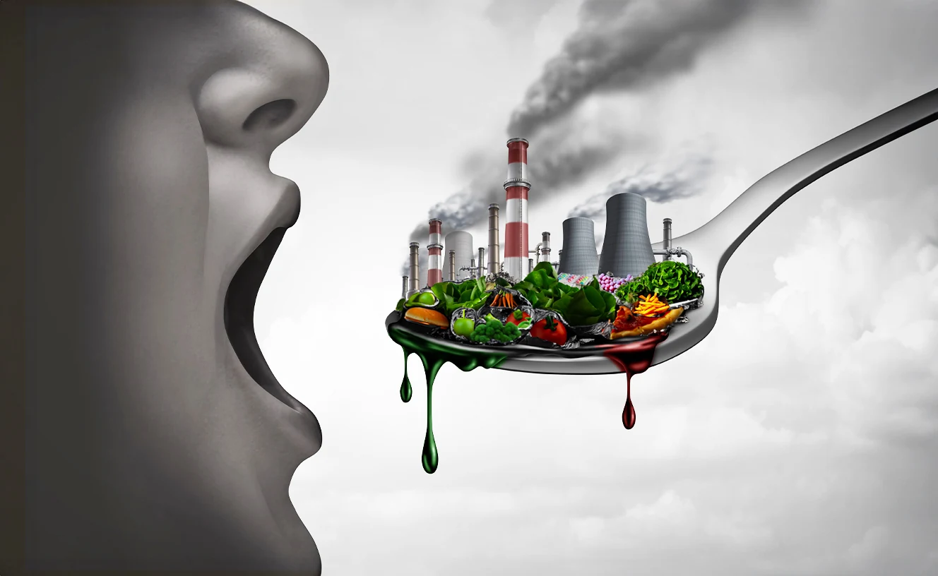 A large gray face in profile with an open mouth faces a spoon that holds factory smokestacks and cooling towers, emitting dark smoke, among vegetables, a tomato and other food, with green and red liquid dripping from the spoon.