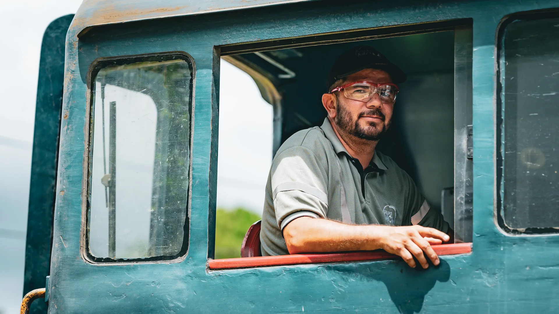 A train driver in glasses and a cap leans out of the cab window of a blue locomotive.