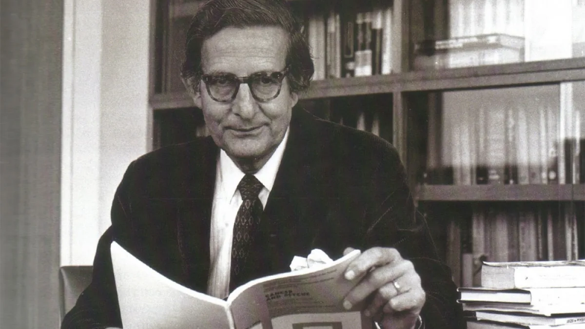 A black and white photograph of a man with dark rimmed glasses, a dark jacket, a white shirt and a patterned tie, smiling slightly and holding an open booklet, with bookshelves behind him and a stack of books at the lower right.