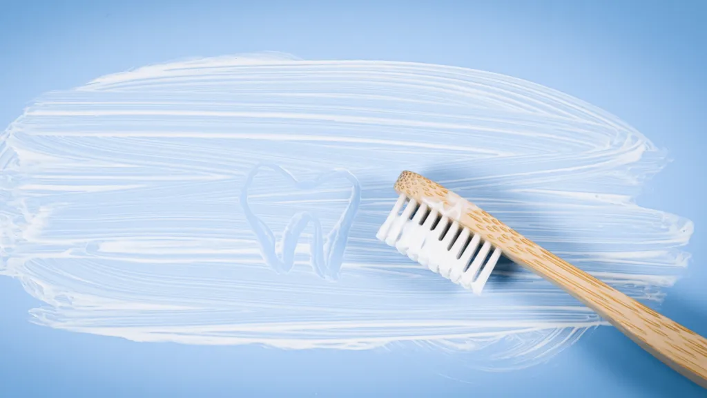 A wooden handled toothbrush lying on a pale blue surface beside a smear of white toothpaste with a tooth shape drawn in it.