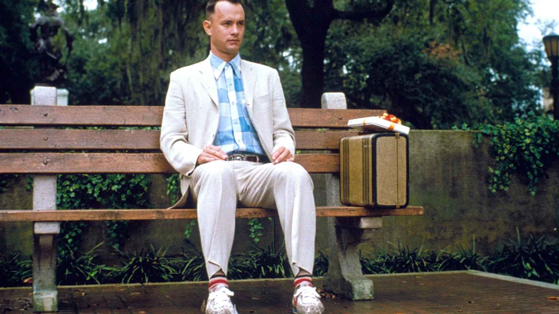 A man in a light linen suit and a blue checked shirt sits on a wooden park bench with a small tan suitcase and a white box tied with an orange ribbon beside him, with trees and a stone wall behind.