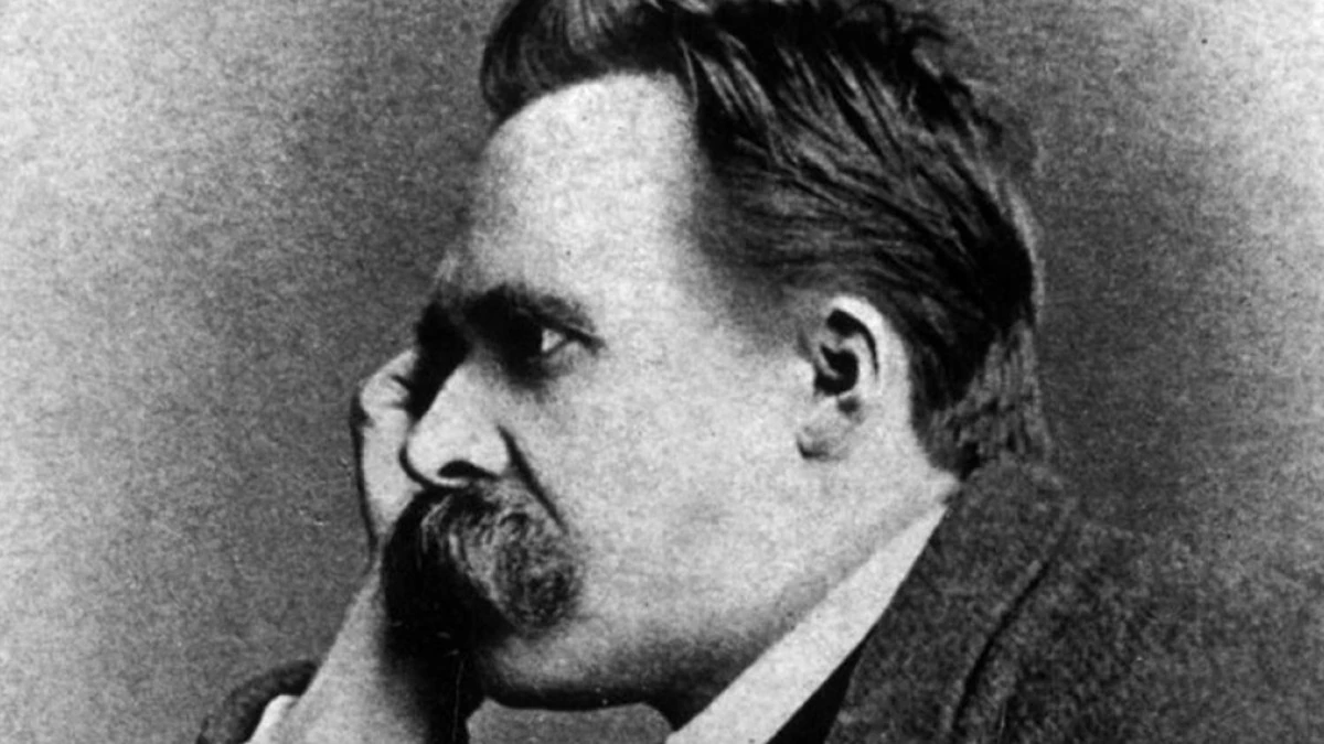 Black and white photograph of a man in profile facing left, with swept back hair and a thick mustache, resting the side of his head against his hand, in a dark coat with a white collar.