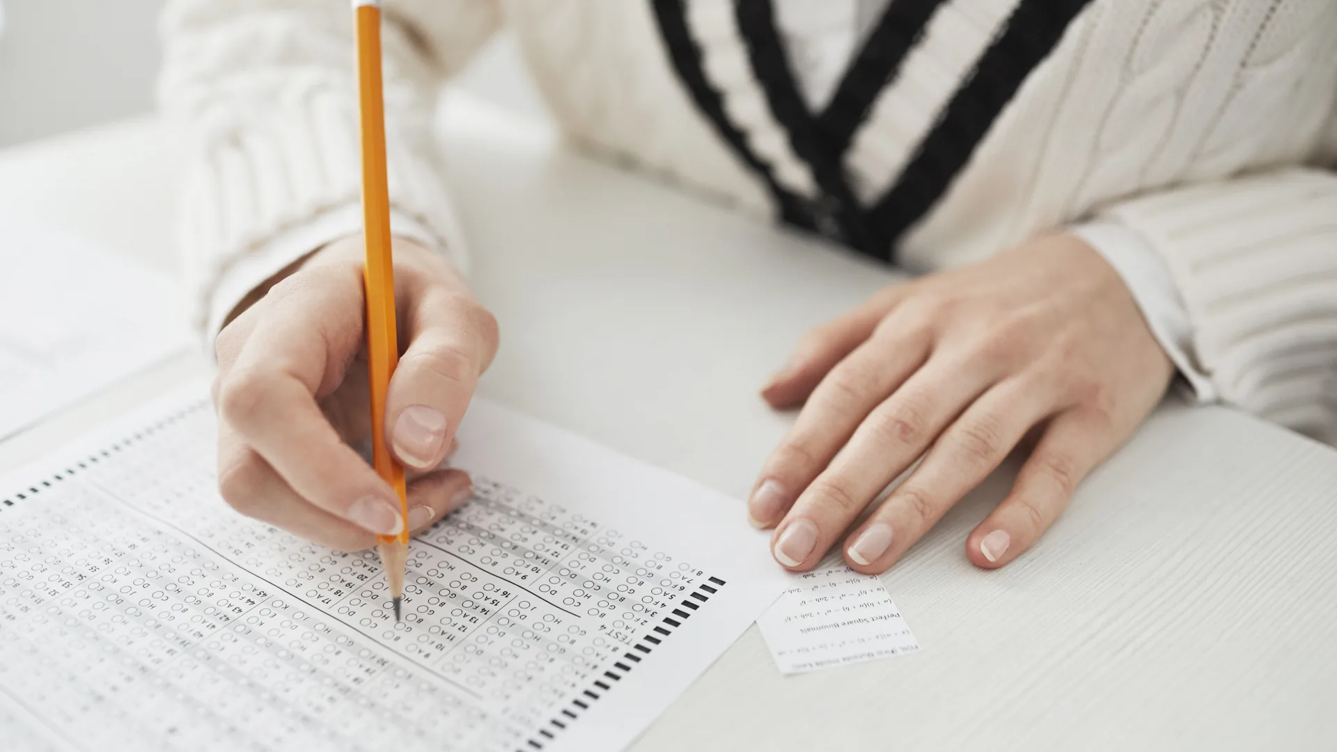 A hand holding a pencil fills in a printed answer sheet on a white desk.