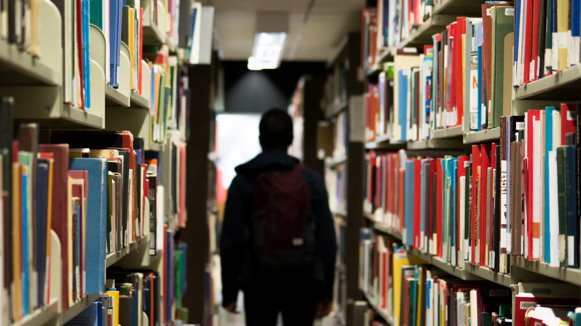A person wearing a backpack walks between tall library shelves filled with books.