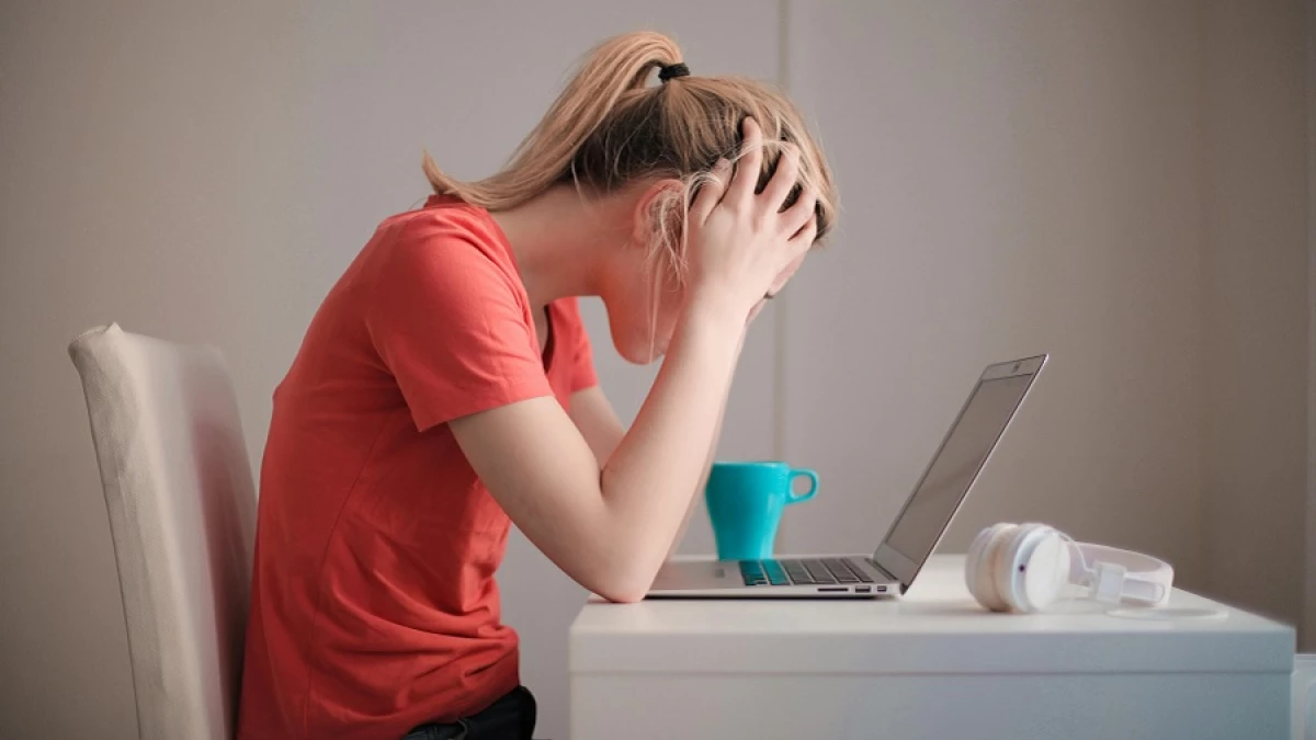 A person in a red shirt sitting at a white desk with a laptop, head in hands, next to a teal mug and white headphones.