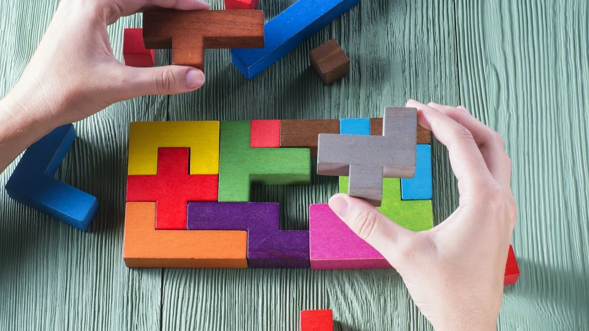 Two hands fitting wooden puzzle pieces of different shapes and colors into a rectangular arrangement on a gray green wooden table, with a few loose pieces lying around the edges.