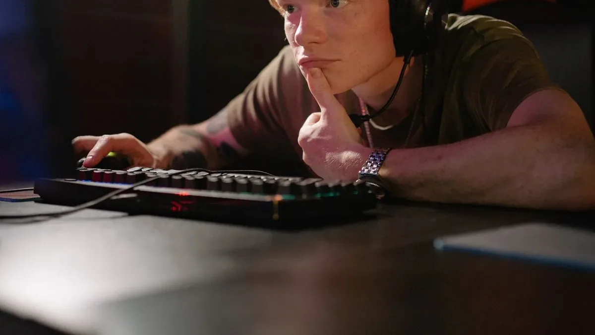 A young man with short light hair and a headset leans on a dark desk with his chin resting on one fist, looking toward a screen out of frame, while his other hand rests on a mouse next to a backlit mechanical keyboard.