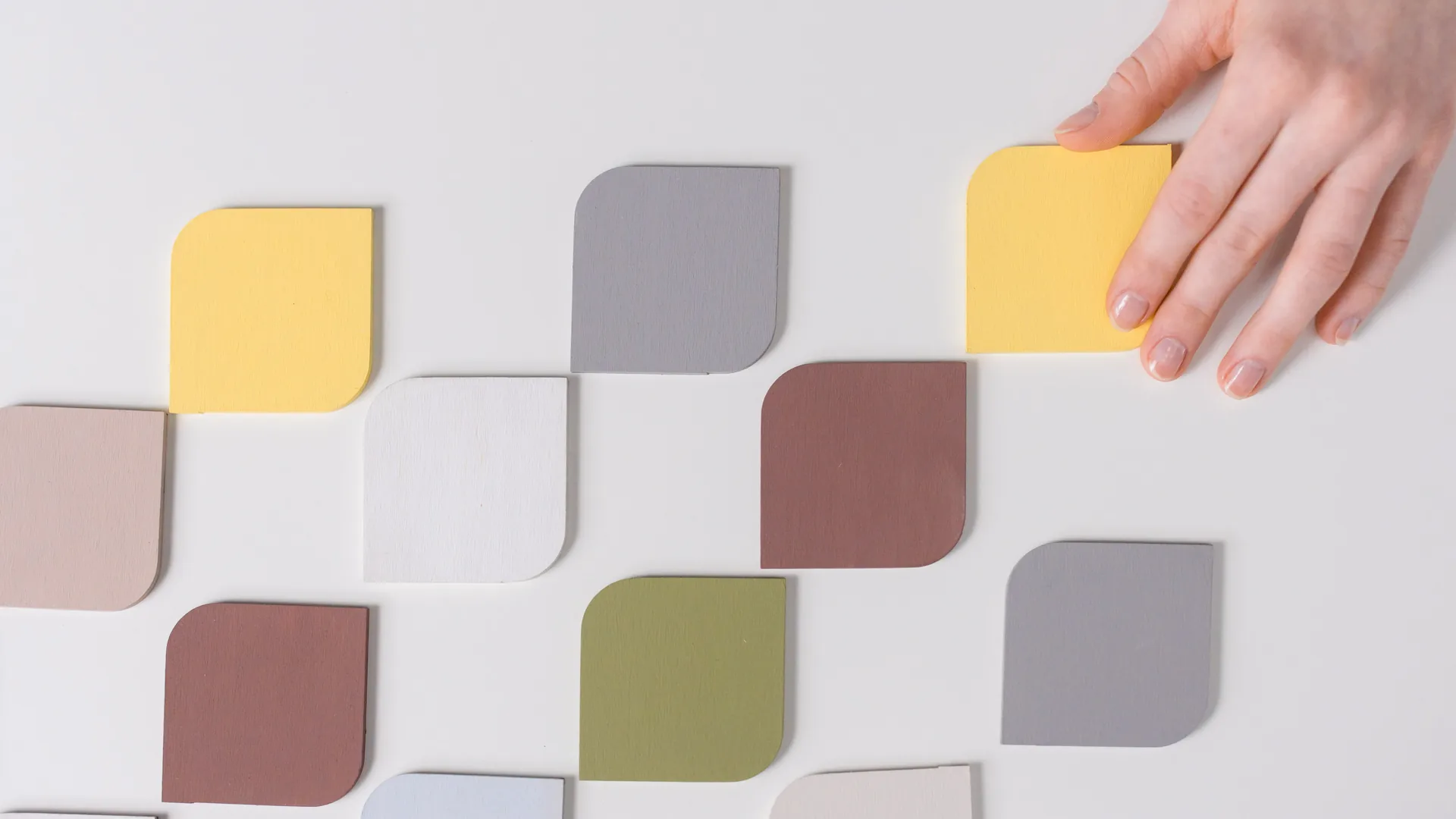 A hand places a yellow tile into a staggered grid of pastel tiles on a white surface.