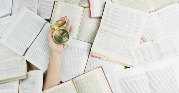 Many open books lie spread across a surface, and a human hand rises from among them holding a small brass pocket compass in its open palm.