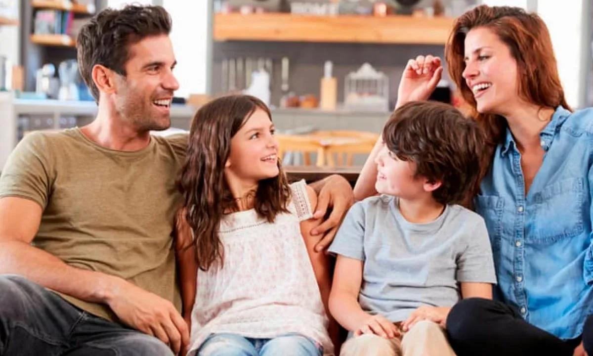 A man in an olive green T-shirt and a woman in a denim shirt sit close together on a couch laughing, with a girl in a white sleeveless top between them and a boy in a gray T-shirt beside the woman, in a bright kitchen with shelves behind them.