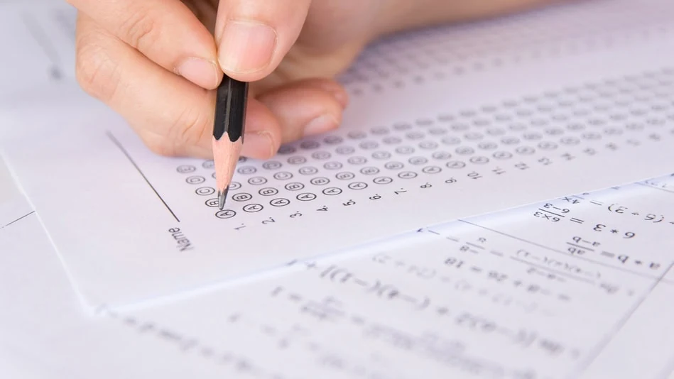 A hand holds a pencil over a printed answer sheet of numbered bubble rows, with sheets of printed algebra problems lying beside and beneath it.