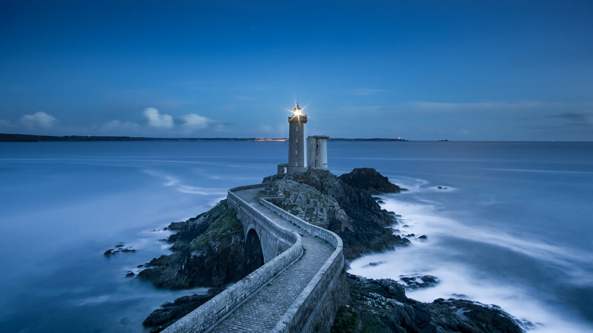 A lighthouse on a rocky coast with a stone walkway leading toward it.