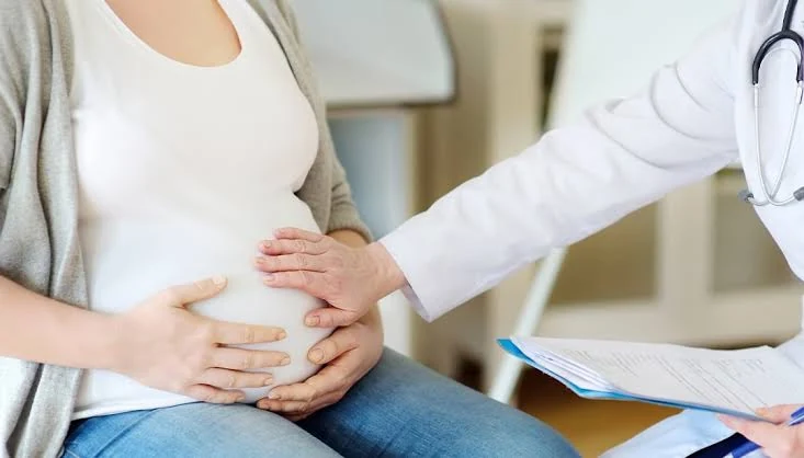 A seated pregnant woman in a cream top and jeans holds her abdomen with both hands while a person in a white coat with a stethoscope rests a hand on it and holds a blue folder of papers.