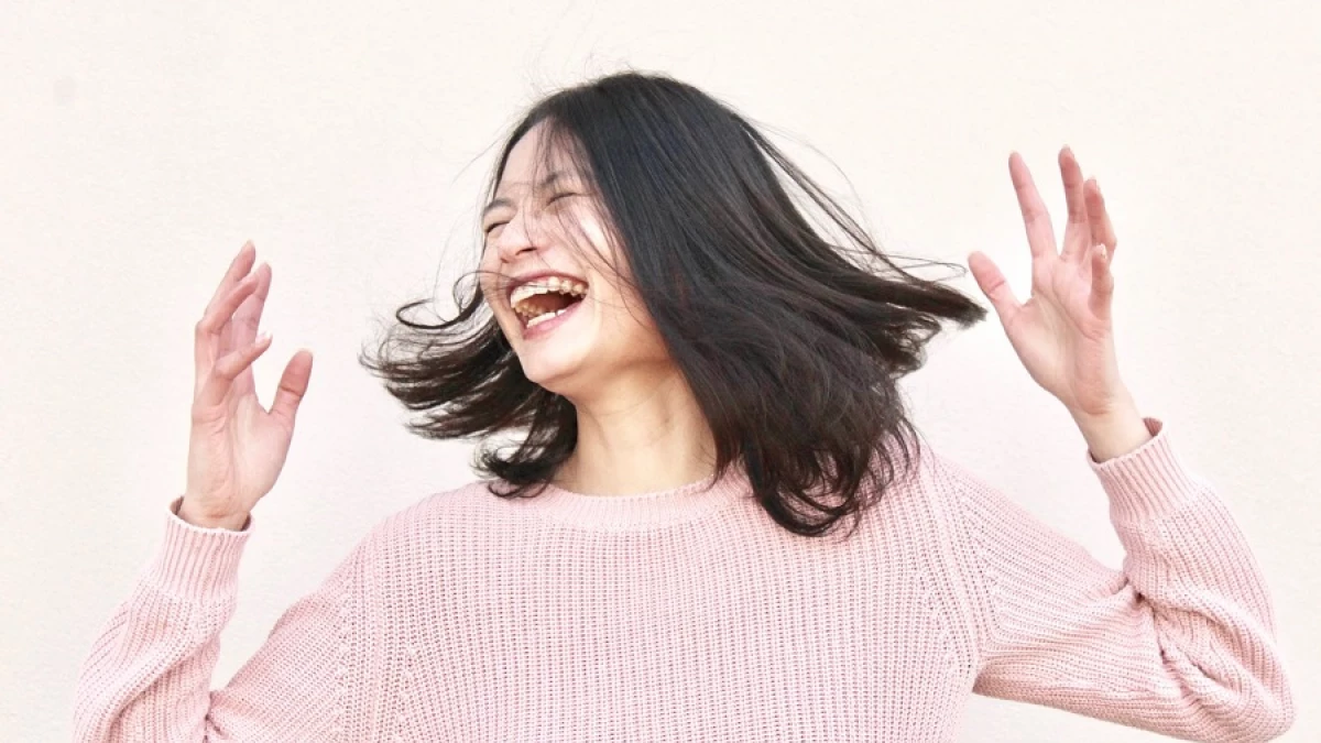 A woman in a pink knit sweater laughing with her eyes closed, hands raised and hair caught mid-motion, against a plain pale background.