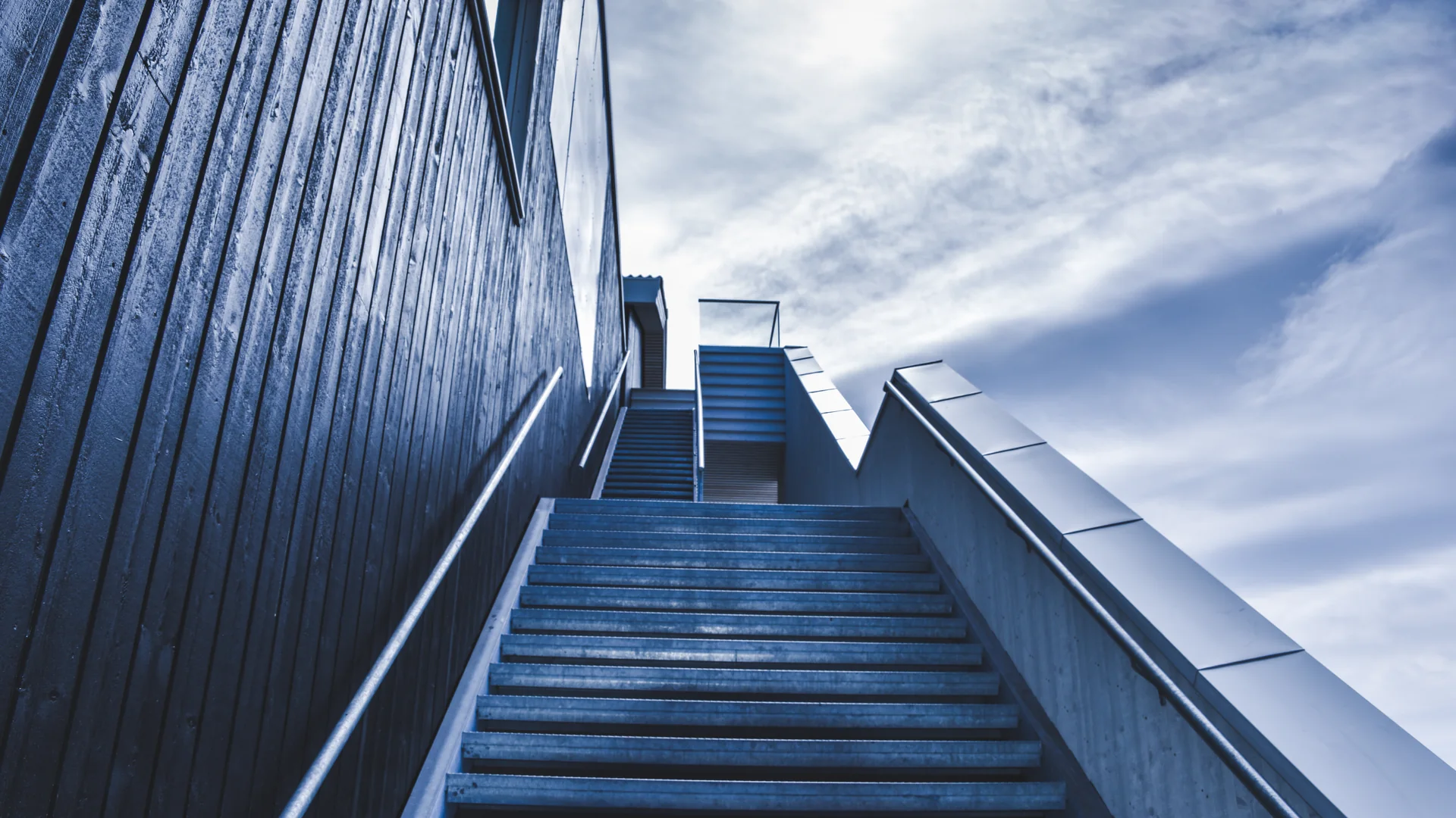 A blue grey outdoor staircase rises beside a dark wooden building, with metal handrails and a cloudy sky above the top landing.