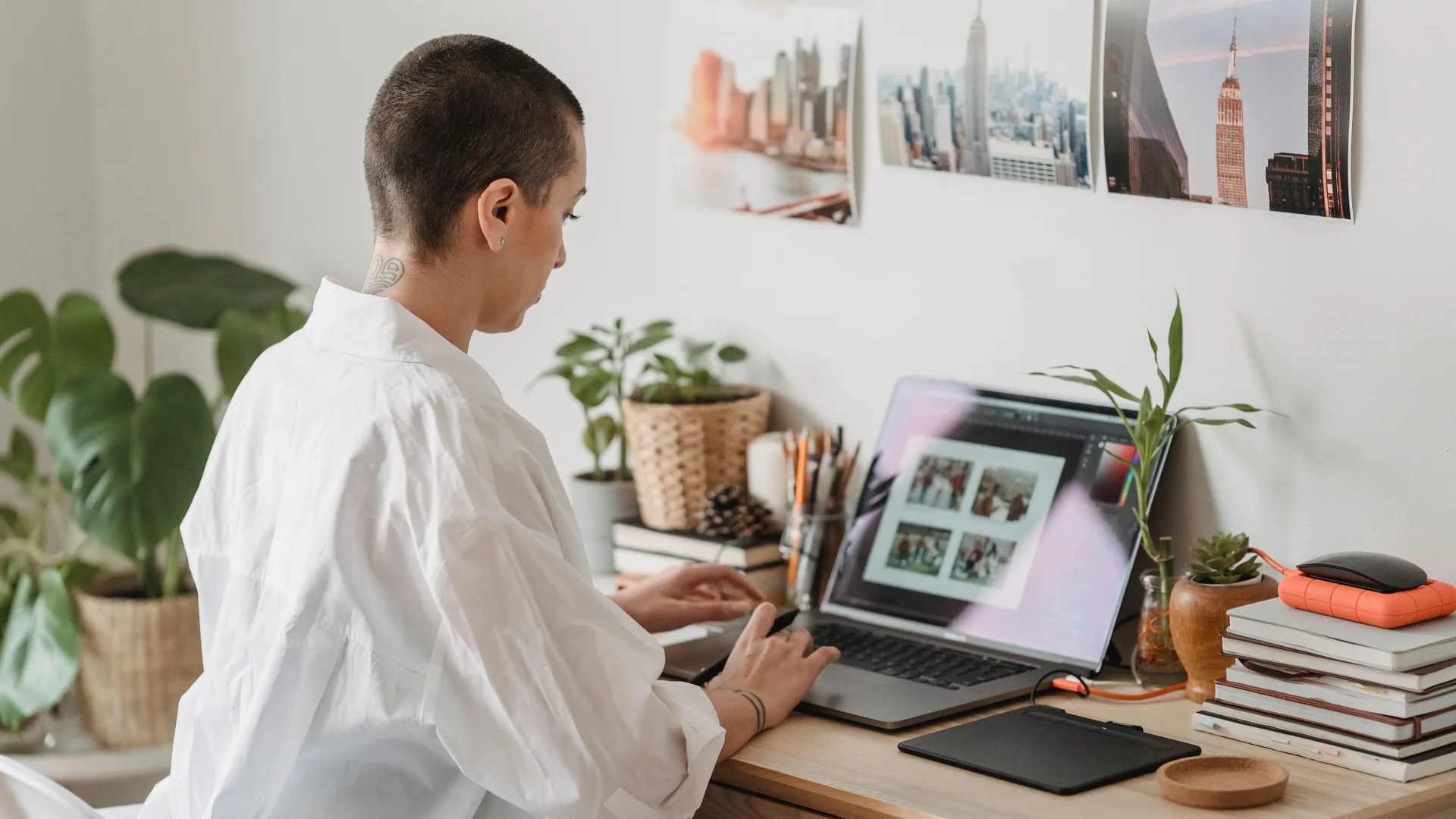 A woman with a buzz cut and a white shirt works at a laptop beside a pen tablet, plants and a stack of notebooks.