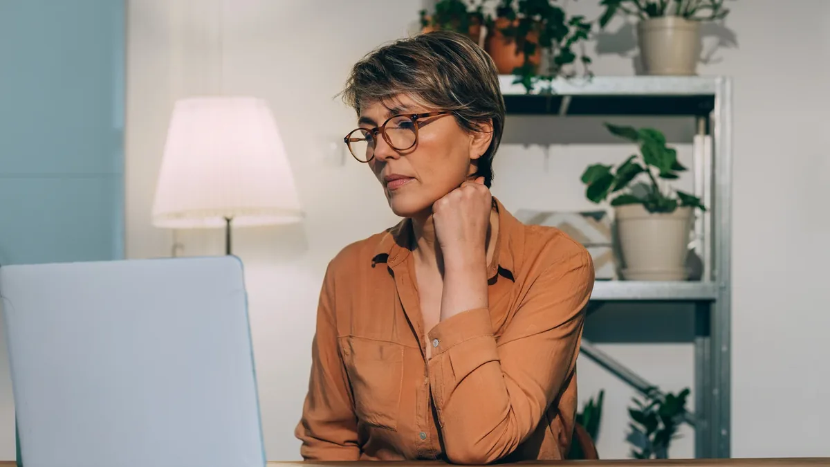 A woman with short hair and glasses sits at a laptop with one hand at her neck, in a room with shelves and plants.
