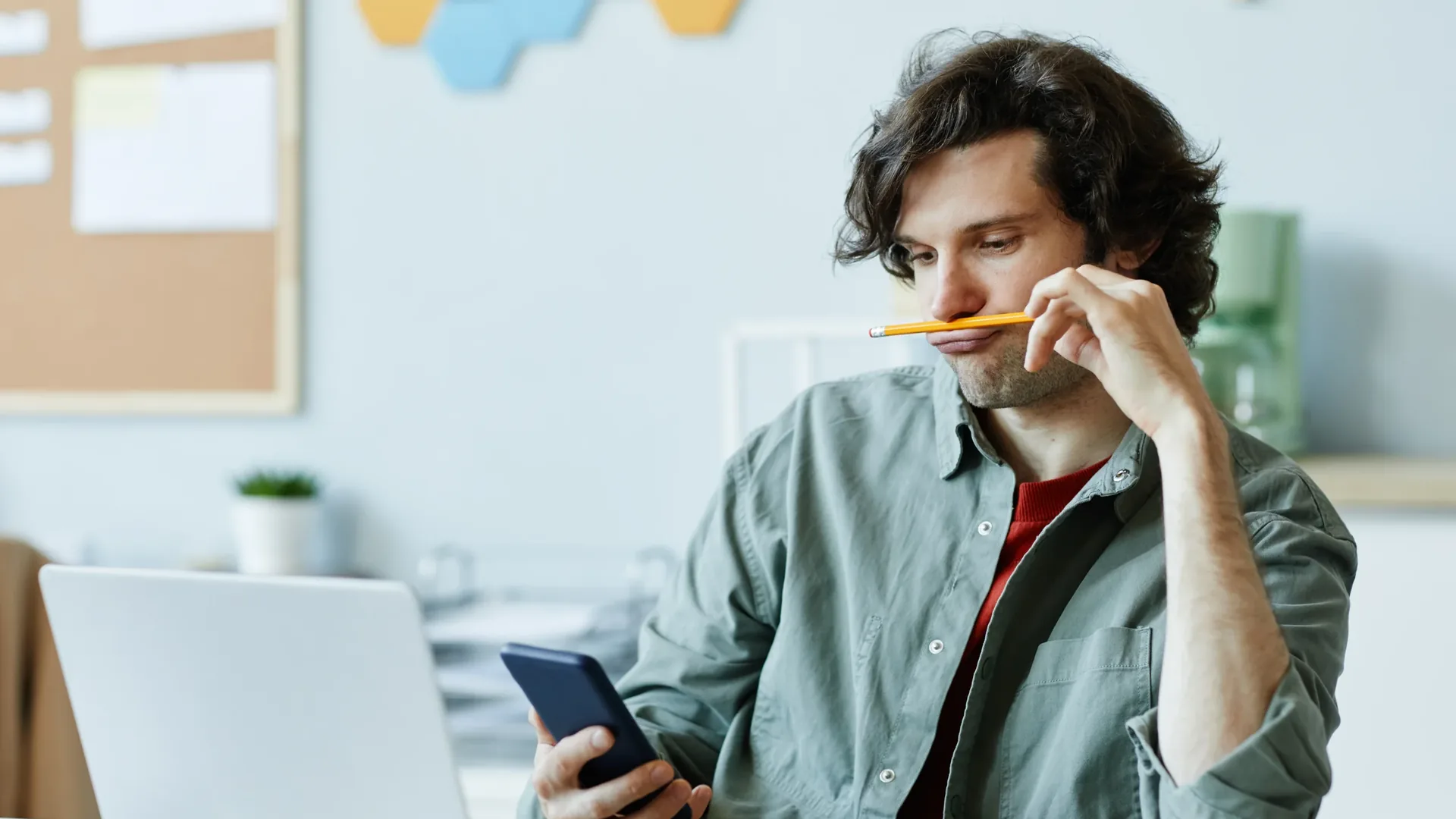 A man with dark wavy hair and stubble sits at a desk with a silver laptop, holding a smartphone in one hand and a yellow pencil under his nose with the other, in a bright office with a cork board and hexagonal wall panels behind him.