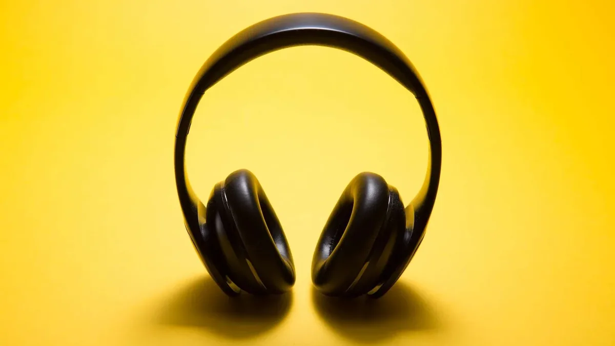 Black over ear headphones standing upright on a bright yellow background, with the curved headband above two cushioned ear cups and soft shadows on the surface below.